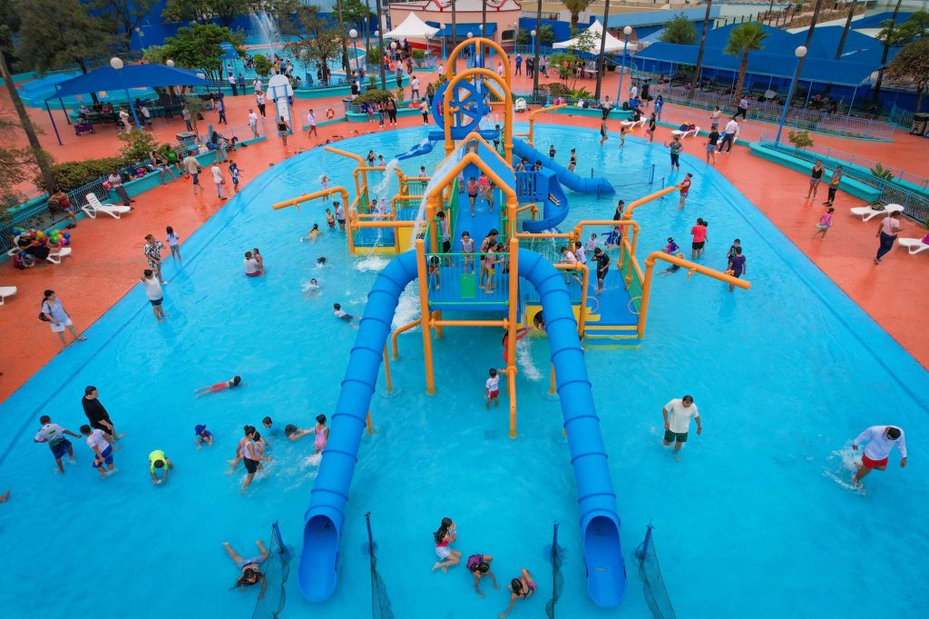 Vista aÃ©rea de alberca infantil con estructura de juegos acuÃ¡ticos, toboganes azules y decenas de familias baÃ±Ã¡ndose.